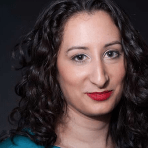 A woman from below shoulders up, with long dark curly hair, olive skin, red lips, brown eyes wearing a turquiose shirt.