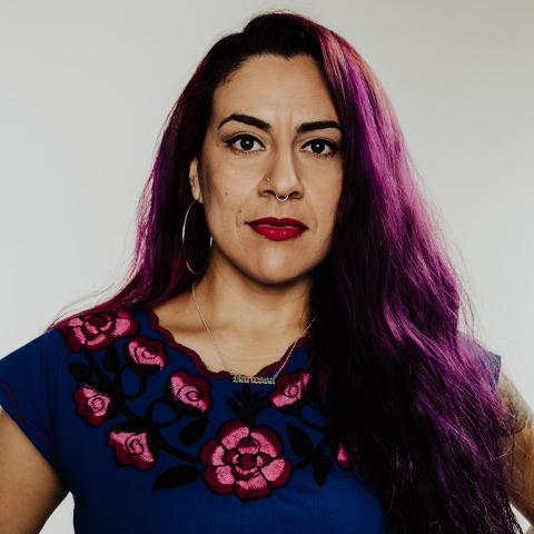 Vanessa has light skin and purple hair. She poses in front of a white backdrop in a floral dress with red lipstick on.