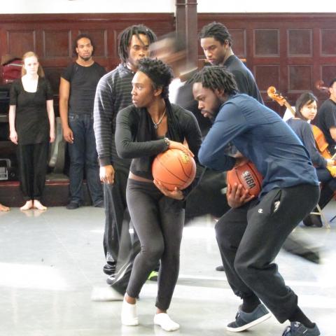 Dancers perform with basketballs in the foreground, while other dancers and instrumentalists look on in the background.