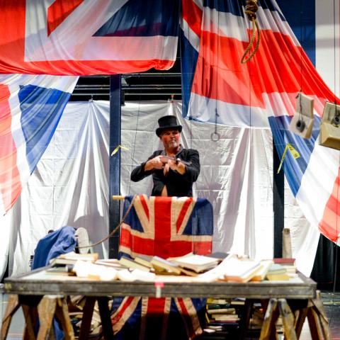 Nora, in a top hat, performs on a stage with British flags.