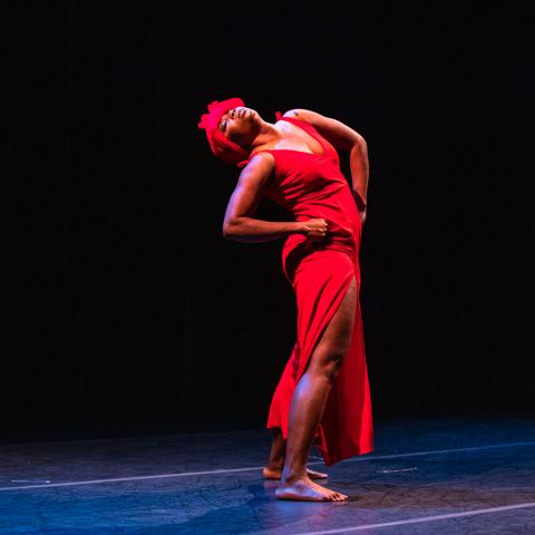 A Black woman, in a red hat and dress, leans back and holds her hips.