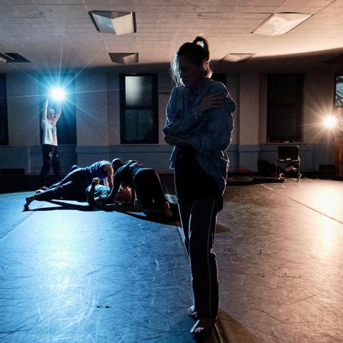 In a room with a low ceiling and dramatic lighting, a woman is in the foreground and she holds herself. In the background, three more folks lay on top of each other.