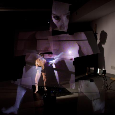 Projections of different parts of a female surround a nude male figure in a dark room.